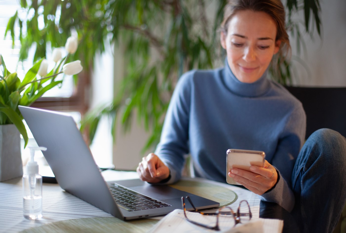Ung kvinne sitter komfortabelt ved et hjemmekontorbord omgitt av grønne planter og jobber på en laptop mens hun holder en smarttelefon. Hun smiler mens hun ser på telefonen, med briller og en notatbok ved siden av seg på bordet.